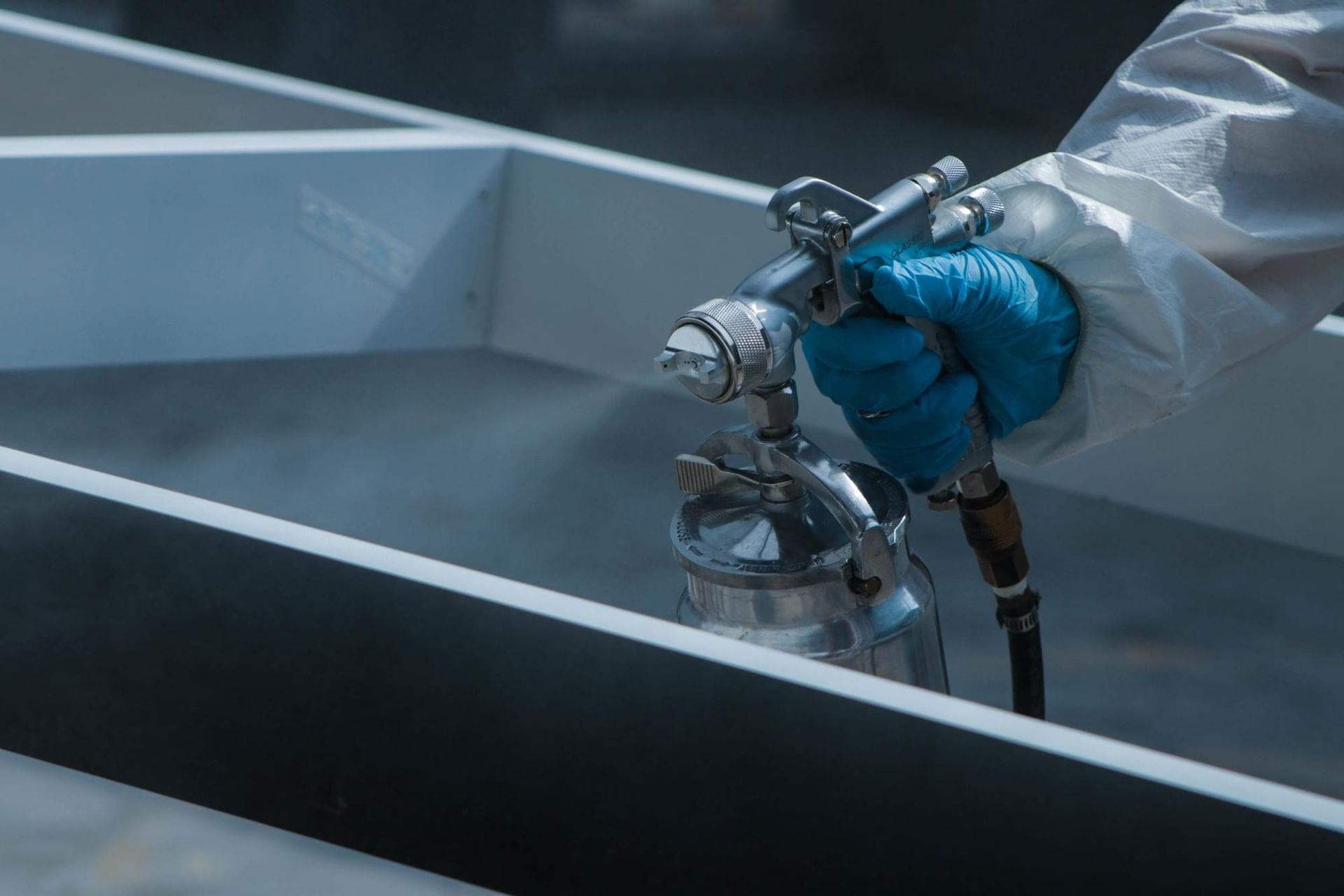 Person in a protective suit and blue glove spraying coating onto a gray metal panel